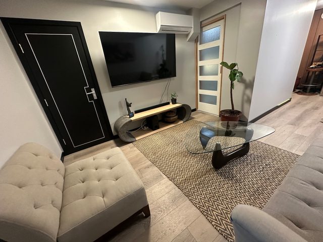 Living room seen from the tufted sofa: wall TV on a curved wood console, glass coffee table, fiddle-leaf fig and chevron rug